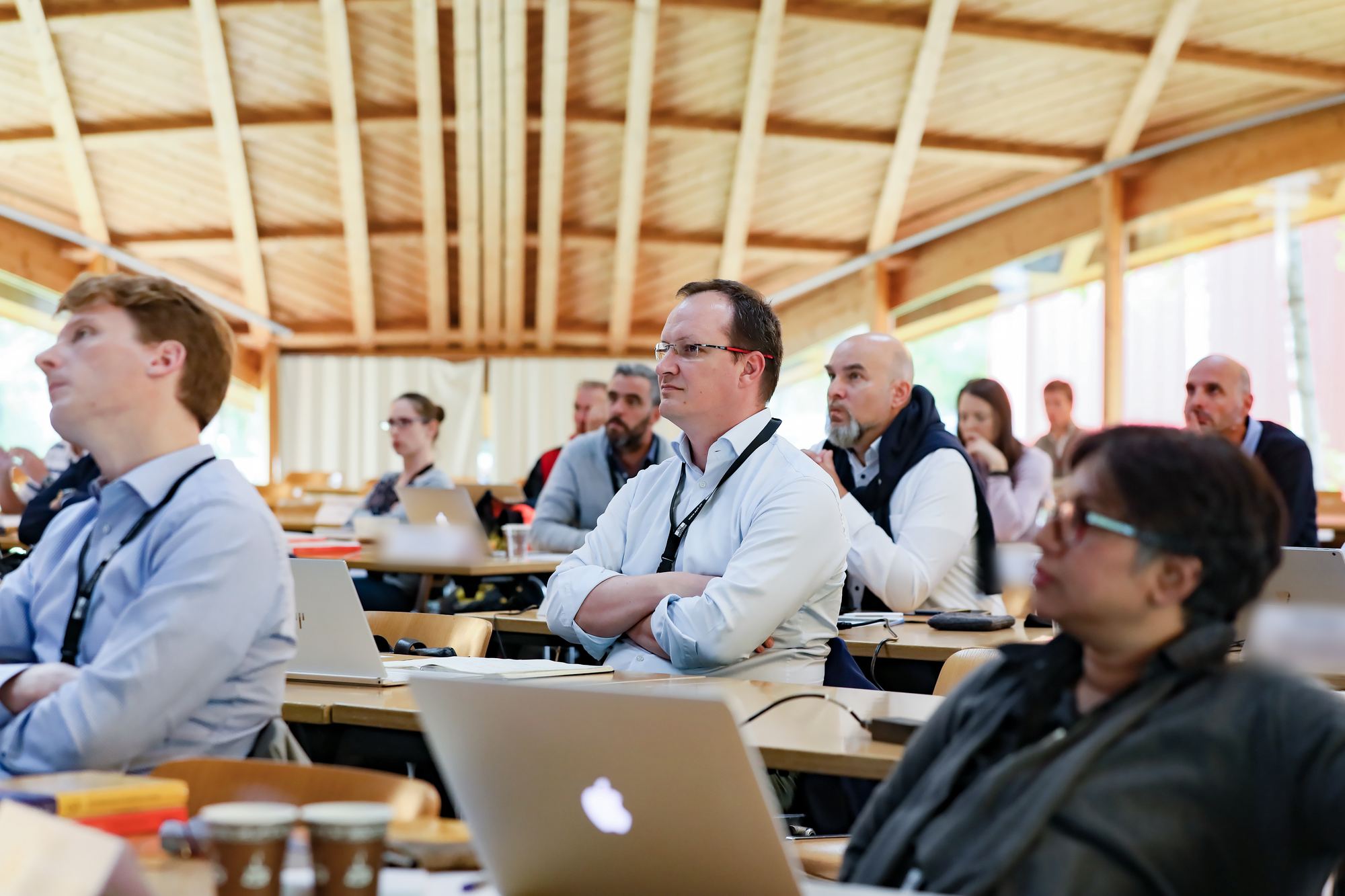 A room of executive-program participants listening attentively, laptops open, under a wooden roof